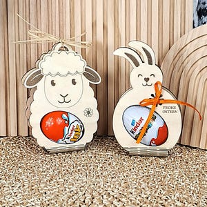 May include: Two wooden Easter decorations: a sheep and a bunny, each holding a Kinder Joy egg. The sheep has a raffia bow, and the bunny has an orange ribbon. The bunny decoration has the words "FROHE OSTERN" on it.