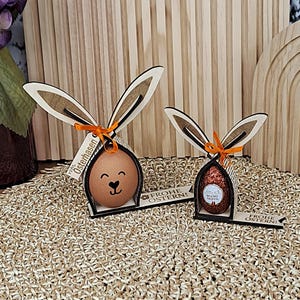 May include: Two Easter bunny-shaped wooden decorations. One holds a brown egg with a smiling face, the other a chocolate egg. Both have wooden ears and orange ribbon. The text "Frohe Ostern!" is visible.