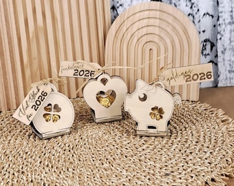 May include: Three wooden decorative boxes in the shape of a clover, heart, and pig, each with a small opening and a tag that reads "2026". The boxes are light brown and sit on a woven mat.