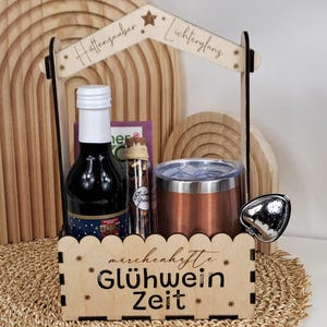 Puede incluir: Un juego de regalo de madera con una botella de vino, un vaso de color cobre y un vial de vidrio de especias. El juego está decorado con un arco de madera y un letrero que dice "Glühwein Zeit". El juego está sobre una estera tejida.
