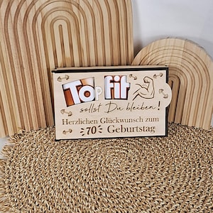 以下が含まれることがあります： 「Top Fit」と「Herzlichen Glückwunsch zum 70. Geburtstag」の文字が刻まれた木製の箱。箱は黒い縁取りがあり、ダンベルと腕のグラフィックで装飾されています。背景には、木製のアーチ型の装飾が含まれています。