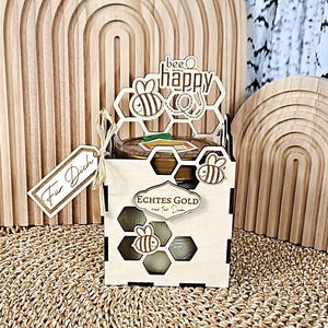Könnte beinhalten: Ein hölzernes Honig-Geschenkset mit einem Honigglas in einer lasergeschnittenen Box. Die Box hat Bienen- und Wabenmuster mit den Worten "bee happy" und "Echtes Gold nur für Dich". Ein kleines Etikett sagt "Für Dich".