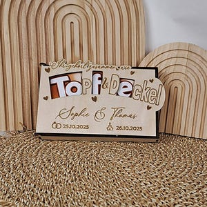 May include: A wooden wedding gift with the words "Topf & Deckel" and the names "Sophie & Thomas" engraved. The dates 25.10.2025 and 26.10.2025 are also included, along with heart and ring symbols.