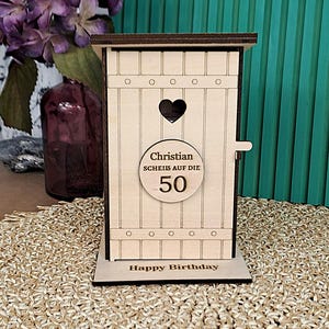 Könnte beinhalten: Eine holzfarbene Geschenkbox in Form eines Toilettenhäuschens mit herzförmigem Ausschnitt und dem Text "Christian Scheiß auf die 50". Auf der Basis steht "Happy Birthday".