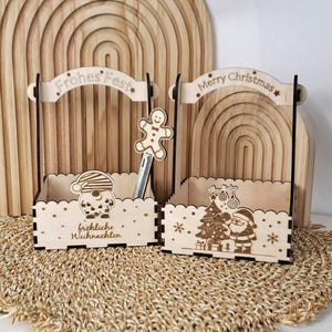 Puede incluir: Dos cestas navideñas de madera cortadas con láser con asas, una con pancartas "Frohes Fest" y la otra "Merry Christmas". Cada cesta presenta diseños festivos, incluyendo un hombre de jengibre, Papá Noel y un árbol de Navidad.