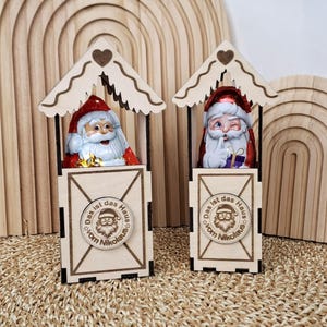 Può includere: Due statuette di Babbo Natale in legno in piccole casette. Ogni Babbo Natale ha un cappello rosso e barba bianca. Le casette hanno un tetto a forma di cuore e un emblema circolare con testo. Lo sfondo include archi in legno.