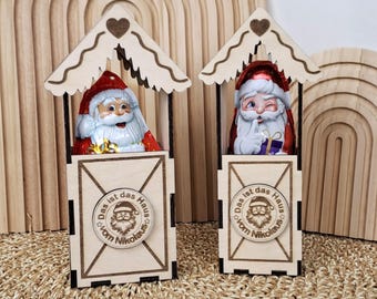 Pode incluir: Duas figuras de madeira do Pai Natal em pequenas casas. Cada Pai Natal tem um chapéu vermelho e barba branca. As casas têm um telhado em forma de coração e um emblema circular com texto. O fundo inclui arcos de madeira.
