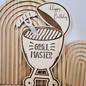 Könnte beinhalten: Holz-Geburtstagsdekoration mit Grill-Design, den Worten "Grill Master" und "Happy Birthday". Die Dekoration enthält einen Spatel und einen Deckel. Der Hintergrund zeigt Holzbögen und eine gewebte Matte.