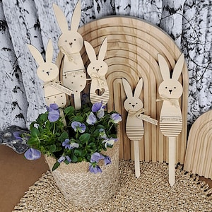 May include: Wooden Easter bunny decorations with attached stakes, some with the text "HAPPY EASTER" and "FROHE OSTERN". The bunnies are arranged around a woven basket of purple flowers, with a wooden rainbow in the background.
