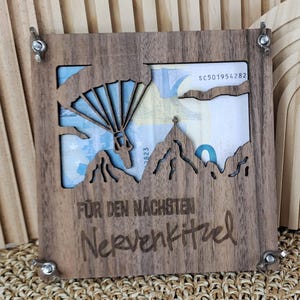 Könnte beinhalten: Ein Geldhalter aus Holz mit einem Berg- und Fallschirmdesign. Der Rahmen ist dunkelbraun und das Design ist ausgeschnitten, um das Geld im Inneren freizulegen. Der Text "Für den nächsten Nervenkitzel" ist unten eingraviert.