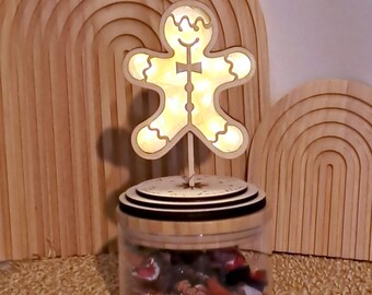 May include: A decorative jar with a wooden gingerbread man topper. The gingerbread man is illuminated with a warm light. The jar is filled with colorful candies and sits on a woven mat. Wooden arch accents are in the background.