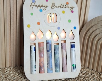 May include: A wooden birthday card with the words "Happy Birthday" and the number "60" cut out. The card has six slots holding rolled-up banknotes, and is decorated with colorful confetti.
