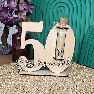 Puede incluir: Centro de mesa de madera con el número '50', un pequeño vial de vidrio con un objeto enrollado y la palabra 'Glückwunsch' en cursiva. El centro de mesa está sobre una superficie tejida, con un arreglo floral morado y un arco turquesa al fondo.