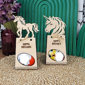 May include: Two wooden gift boxes with unicorn toppers and the words "Happy Birthday." Each box contains a chocolate egg. The background features a green arch and a woven mat.