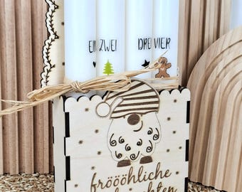 May include: A wooden box with a gnome design holds four white candles. The candles are labeled with numbers and festive illustrations. The box has the words "fröhliche Weihnachten" and star cutouts. The background features wooden arches.
