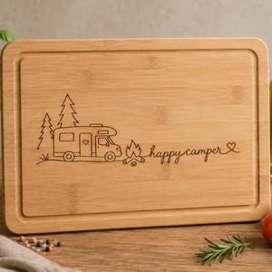 Op de afbeelding: Een rechthoekige snijplank van bamboe met een lasergegraveerd ontwerp van een camper, dennenbomen, een kampvuur en de tekst "happy camper" met een klein hartje. De plank is voorzien van een groef langs de rand van het bovenoppervlak.