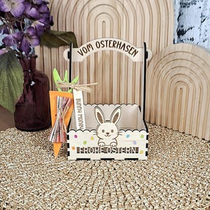 Könnte beinhalten: Ein hölzerner Osterkorb mit Hasenmotiv und den Worten "Vom Osterhasen" und "Frohe Ostern". Der Korb ist mit bunten Punkten verziert und enthält eine Karotte mit einem "Bunny Money" -Etikett.