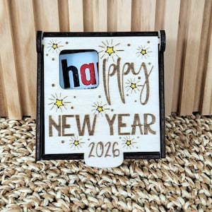 Op de afbeelding: Een houten Nieuwjaarsdecoratie met de woorden "Happy New Year 2026" in een decoratief lettertype. Het woord "ha" is zichtbaar door een klein venster. Het ontwerp bevat steraccenten en een donkerbruin frame.