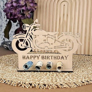 Puede incluir: Un soporte de regalo de cumpleaños con forma de motocicleta de madera con las palabras "HAPPY BIRTHDAY" grabadas en la base. El soporte tiene ranuras para billetes enrollados. El diseño de la motocicleta está recortado de la madera.