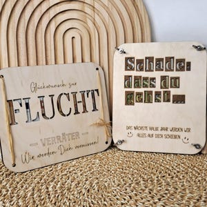 May include: Two wooden plaques with German text. One says "Glückwunsch zur FLUCHT - VERRÄTER - Wir werden Dich vermissen!" The other says "Schade dass du gehst..." and "DAS NÄCHSTE HALBE JAHR WERDEN WIR ALLES AUF DICH SCHIEBEN".