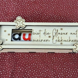 Pode incluir: Uma caixa de madeira com tema natalino com recortes de boneco de gengibre e as palavras "du bist die Glasur auf meinen Lebkuchen!". A caixa tem um painel deslizante com as letras "du".