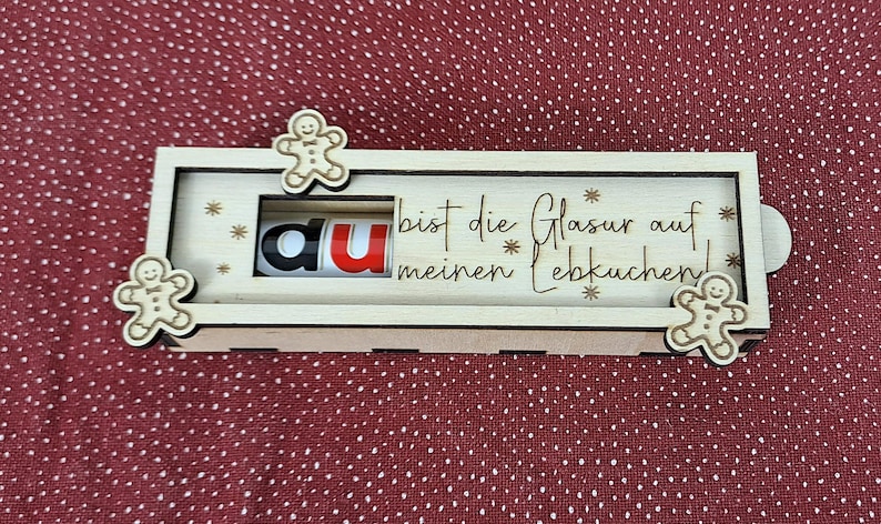 Pode incluir: Uma caixa de madeira com tema natalino com recortes de boneco de gengibre e as palavras "du bist die Glasur auf meinen Lebkuchen!". A caixa tem um painel deslizante com as letras "du".