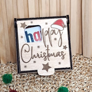 Puede incluir: Una caja de regalo de Navidad de madera con las palabras "Happy Christmas" en una fuente de escritura. La caja tiene una pequeña ventana con la palabra "ha" visible. Un gráfico de gorro de Papá Noel está en la parte superior derecha. La caja está decorada con estrellas y copos de nieve.