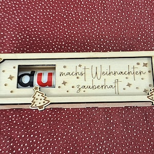 Pode incluir: Uma caixa de madeira com tema natalino com a frase alem&atilde; "du machst Weihnachten zauberhaft" gravada. As letras "du" s&atilde;o vis&iacute;veis numa janela, com o "u" em vermelho. A caixa &eacute; decorada com pequenas &aacute;rvores de Natal de madeira.