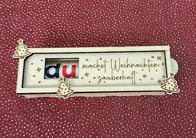 Pode incluir: Uma caixa de madeira com tema natalino com a frase alem&atilde; "du machst Weihnachten zauberhaft" gravada. As letras "du" s&atilde;o vis&iacute;veis numa janela, com o "u" em vermelho. A caixa &eacute; decorada com pequenas &aacute;rvores de Natal de madeira.