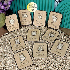 Könnte beinhalten: Eine Sammlung lasergeschnittener Holz-Geschenkanhänger mit verschiedenen Designs, darunter ein Pilz, ein Marienkäfer, ein Schwein und ein vierblättriges Kleeblatt. Jeder Anhänger hat deutschen Text und einen kleinen Holzanhänger.