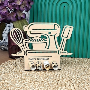 Könnte beinhalten: Ein Geburtstagskartenhalter aus Holz mit Küchenmotiv. Das Design umfasst einen Mixer, einen Schneebesen und einen Spatel. Auf der Basis steht "HAPPY BIRTHAY" und sie hält aufgerollte Nachrichtenrollen. Der Hintergrund zeigt ein türkisfarbenes Bogendesign und lila Blumen.