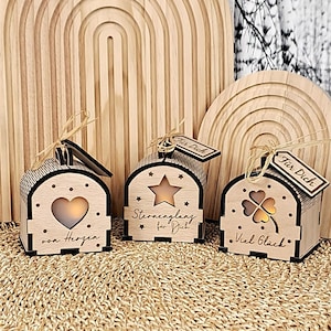 Puede incluir: Tres faroles decorativos de madera con formas recortadas de corazón, estrella y trébol de cuatro hojas. Cada farol tiene una pequeña etiqueta que dice "Für Dich". Los faroles están sobre una estera tejida con un arco de madera en el fondo.