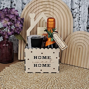 May include: A wooden gift basket with the words "HOME sweet HOME" cut out. The basket contains a bottle of Prosecco, a small candle, and a gift tag. Decorative wooden arches and a vase of purple flowers are in the background.