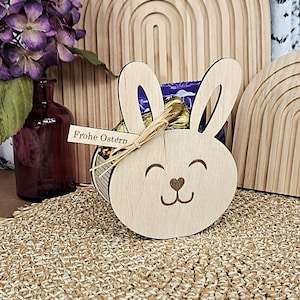 Może przedstawiać: Drewniany pojemnik w kształcie zajączka wielkanocnego z uśmiechniętą twarzą i noskiem w kształcie serca. Pojemnik zawiera zapakowane czekoladki i jest związany rafią. Mała etykieta głosi "Frohe Ostern".