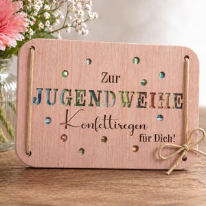 May include: A light pink wooden plaque with the words "Zur Jugendweihe Konfettiregen für Dich!" cut out. The plaque has small circular cutouts and is decorated with a twine bow. A vase of pink flowers is in the background.