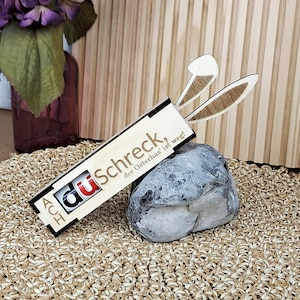 Può includere: Una scatola di legno con la scritta "du Schreck, der Osterhas' ist weg!" e un logo. La scatola ha un paio di orecchie di coniglio di legno che si estendono da essa. La scatola è appoggiata su una pietra grigia.