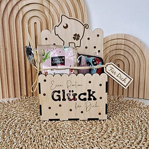 Könnte beinhalten: Eine Holzkiste mit Schweinchen-Ausschnitt und der Aufschrift "Eine Portion Glück für Dich". Die Kiste enthält Tee, eine kleine Tasse und ein Etikett mit "Für Dich". Die Kiste ist mit einer Raffiabastschleife gebunden.