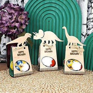 May include: Three wooden dinosaur-themed gift bags with the text "Happy Birthday". Each bag has a different dinosaur cutout on top and contains a colorful egg-shaped treat. The bags are tied with twine.