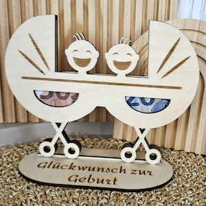 May include: A wooden baby carriage-shaped money holder with two smiling baby faces. The carriage has two compartments for money and the base reads "Glückwunsch zur Geburt". The item is made of light-colored wood.