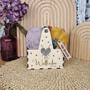 Può includere: Un cestino da lavoro a maglia in legno con un design a cuore e ago, contenente gomitoli di lana lavanda e gialla, ferri da maglia e un segnapunti blu e arancione. Il cestino ha il testo "Wolliebe TO GO" e un'etichetta con la scritta "von Herzen".