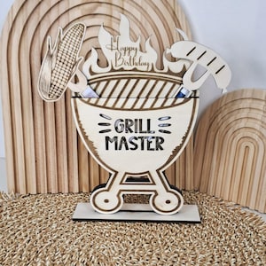 May include: Wooden Grill Master birthday decoration. The design features a barbecue grill with flames, a corn on the cob, and a sausage. The words "Happy Birthday" and "Grill Master" are visible. The decoration is made of light-colored wood.