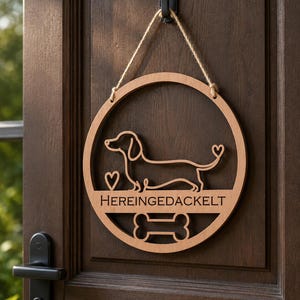 May include: A circular wooden wall sign featuring a laser-cut outline of a dachshund dog, a small heart, and a bone shape. The text HEREINGEDACKELT is engraved across the center. The sign is suspended by a thin jute twine cord from a black hook.