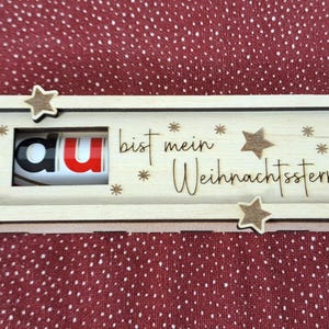 Pode incluir: Uma caixa de madeira com as palavras "du bist mein Weihnachtsstern" e decora&ccedil;&otilde;es de estrelas. A caixa tem um mecanismo deslizante e cont&eacute;m um doce com as letras "au" vis&iacute;veis. O fundo &eacute; um tecido vermelho com pontos.