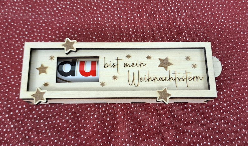 Pode incluir: Uma caixa de madeira com as palavras "du bist mein Weihnachtsstern" e decora&ccedil;&otilde;es de estrelas. A caixa tem um mecanismo deslizante e cont&eacute;m um doce com as letras "au" vis&iacute;veis. O fundo &eacute; um tecido vermelho com pontos.