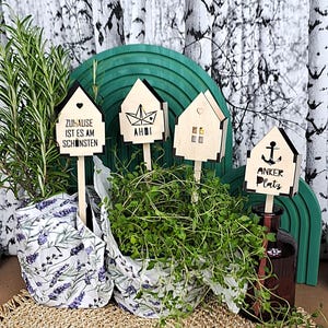 May include: Four house-shaped wooden plant markers on sticks. The first reads ZUHAUSE IST ES AM SCHÖNSTEN with a heart. The second shows a paper boat and AHOI. The third has a window and heart. The fourth features an anchor and ANKER PLATZ.