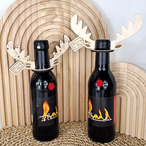 Puede incluir: Dos botellas de vino negro con adornos de cuernos de reno de madera y un pompón rojo. Cada botella tiene una etiqueta con un diseño de llama y la palabra "Flammchen". Los cuernos tienen una etiqueta que dice "Merry X-Mas".