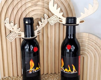 Pode incluir: Duas garrafas de vinho pretas com decorações de chifres de rena de madeira e um pompom vermelho. Cada garrafa tem um rótulo com um desenho de chama e a palavra "Flammchen". Os chifres têm uma etiqueta com "Merry X-Mas".