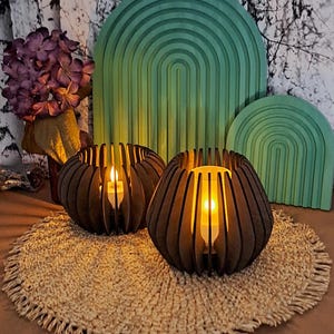 May include: Two brown wooden candle holders with lit candles inside, set on a woven placemat. The candle holders have a unique, slotted design. In the background are green arch-shaped wooden decorations and a bouquet of purple flowers.