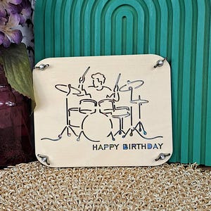 May include: A light-coloured wooden plaque featuring a laser-cut design of a drummer playing a drum kit. The words "HAPPY BIRTHDAY" are below the kit. The square plaque has metal corner accents.