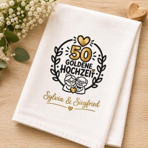 Puede incluir: Toalla de cocina blanca doblada con un diseño dorado y negro que celebra un 50 aniversario de bodas. El diseño incluye las palabras "50 Goldene Hochzeit", un corazón y los nombres "Sylvia & Siegfried".